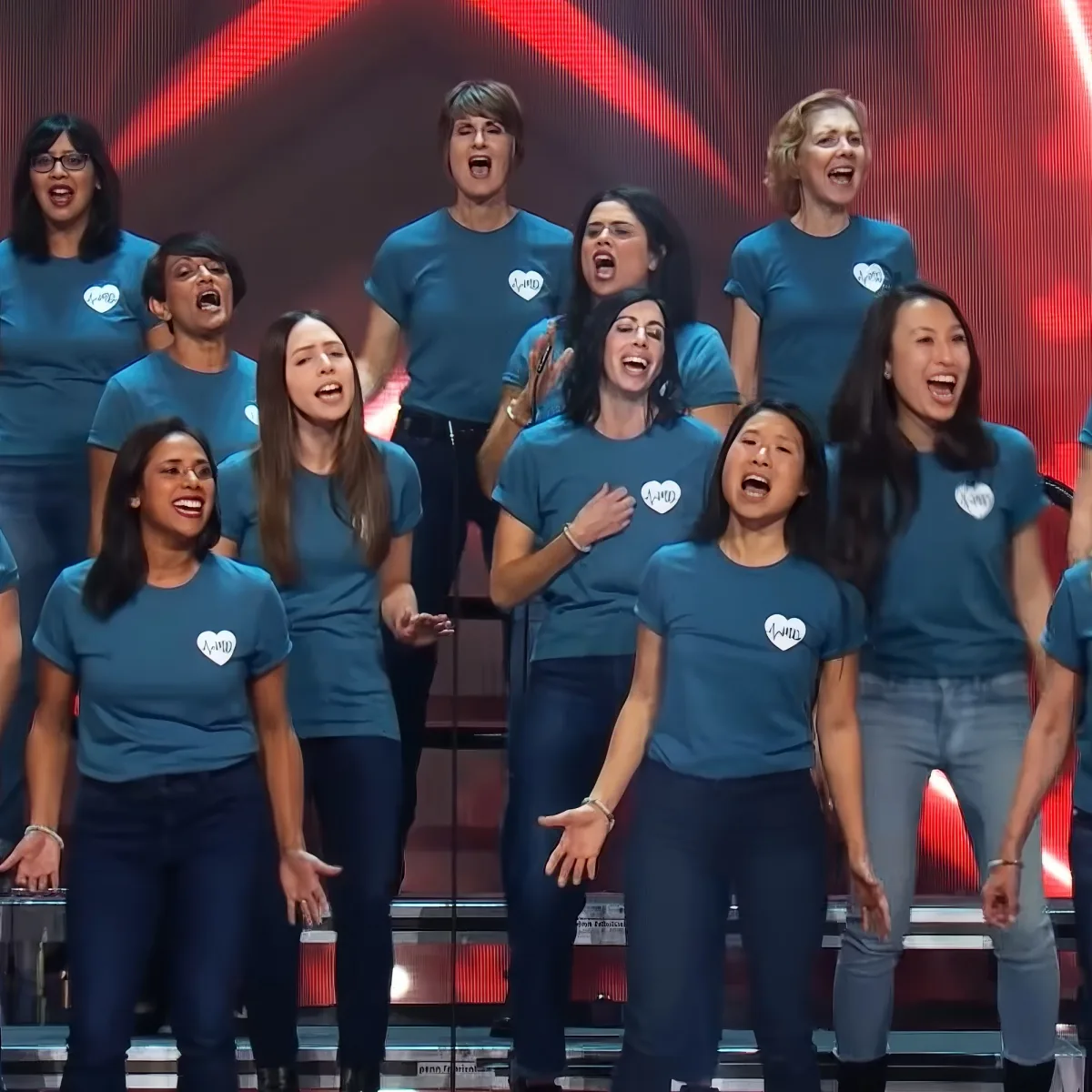 A group of women in blue shirts with hearts sing on stage with red lights behind them.