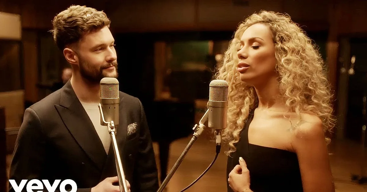 A man and woman are singing in a recording studio, each with a microphone in front of them.