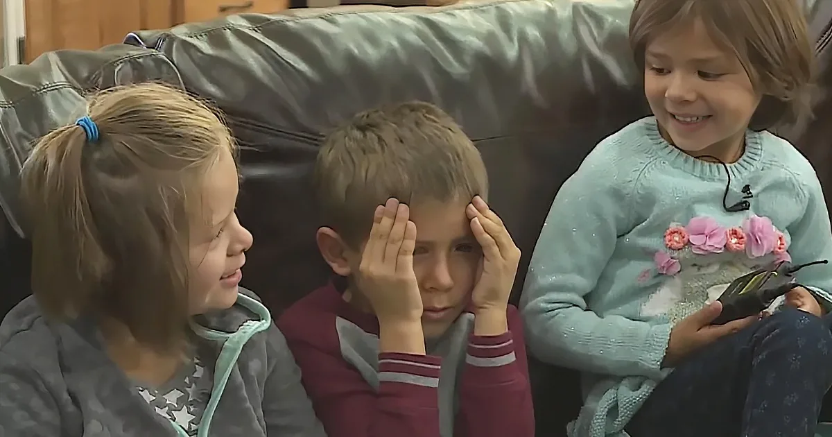 A boy with hands on his face sits between two smiling girls on a couch.