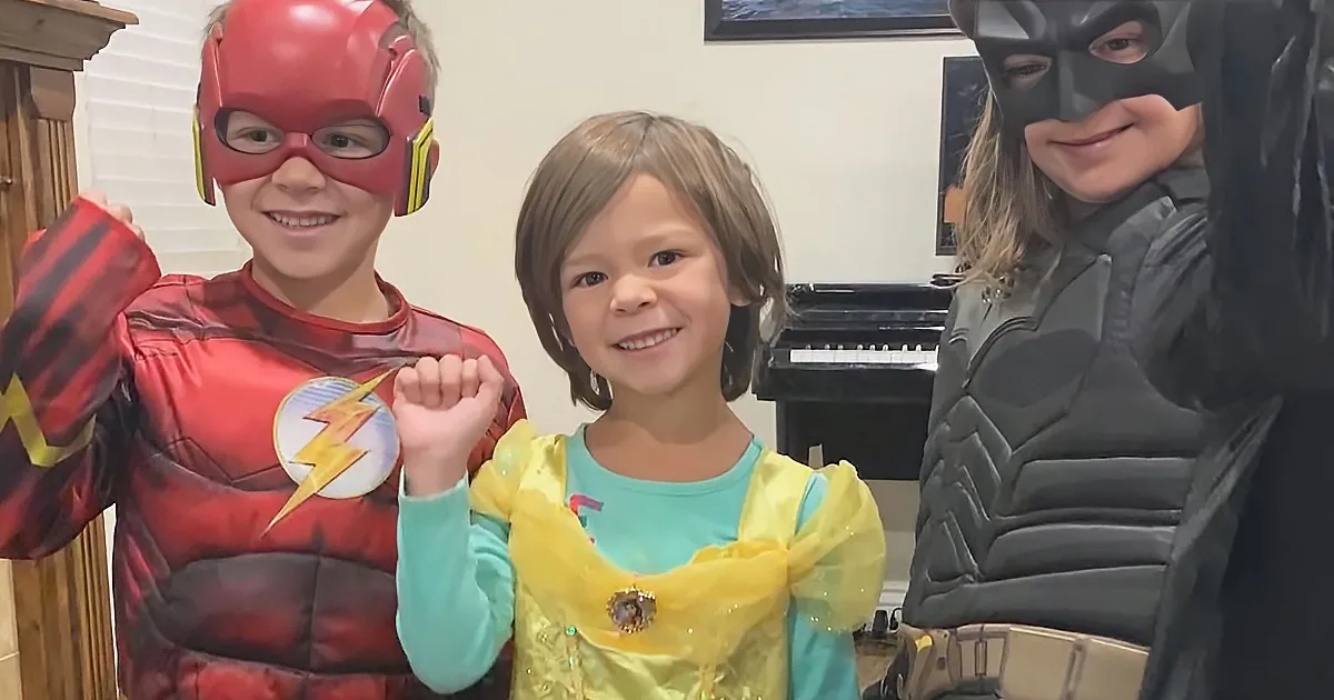 A child in a yellow princess dress stands between a boy in a red superhero costume and a girl in a black superhero costume.