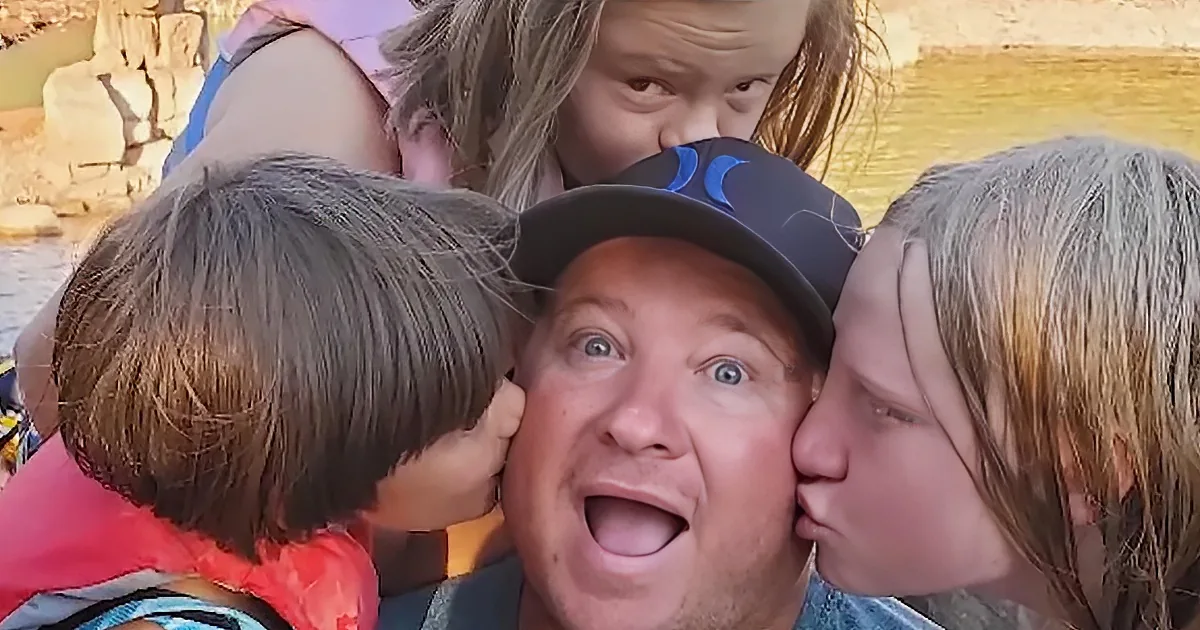 A man wearing a cap is being kissed on both cheeks by two children, with a rocky background and water visible.