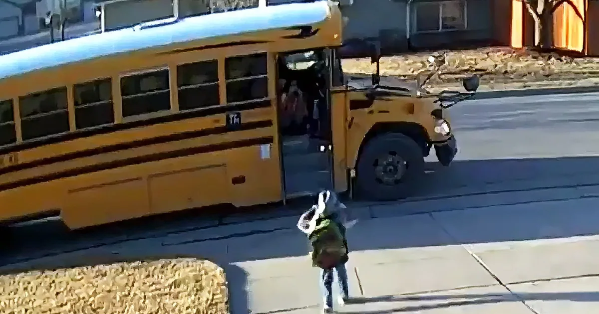 A child in a backpack waves goodbye to a school bus as it drives away.