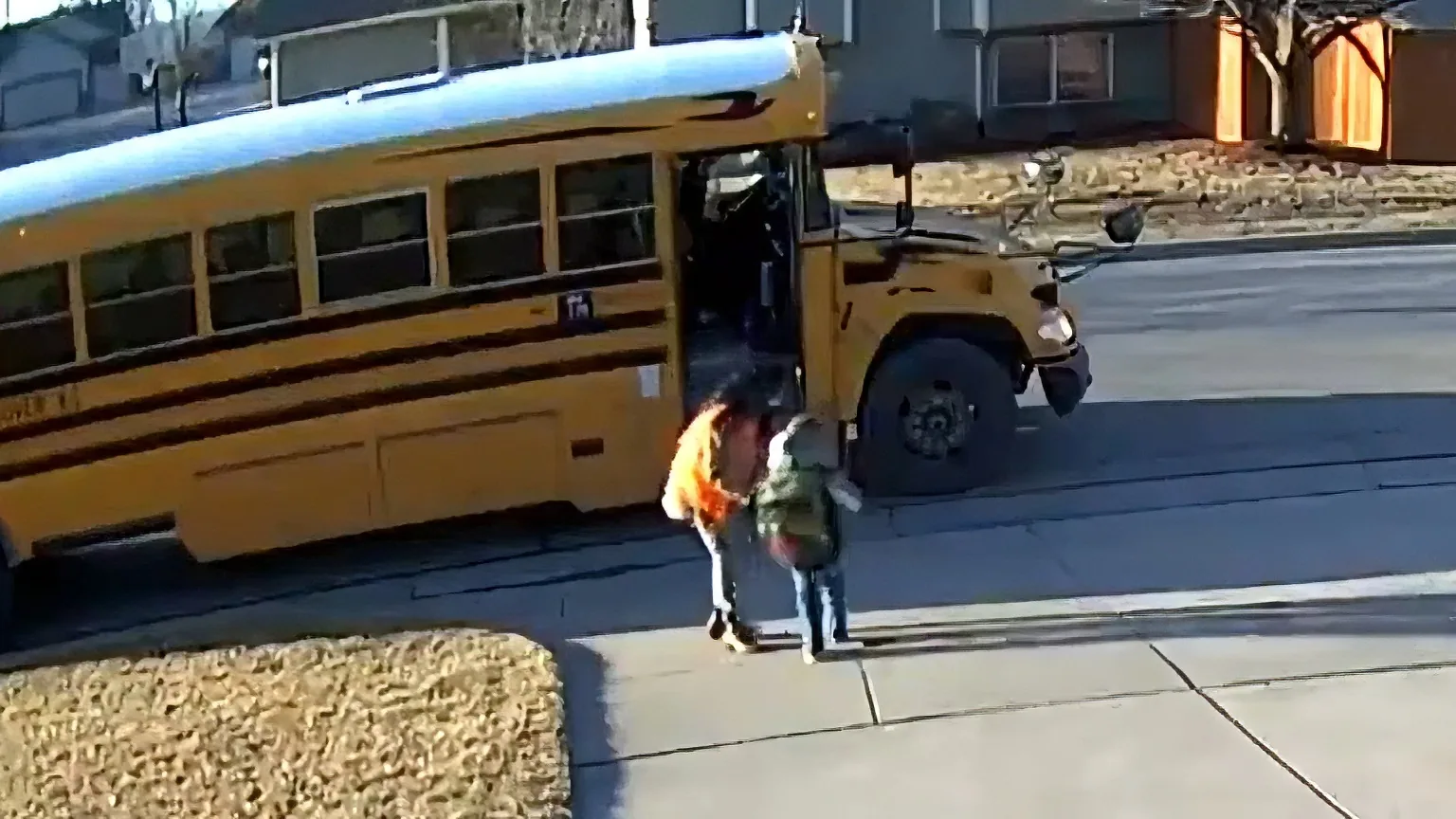 A school bus with its door open, children disembarking, and a dog walking on the sidewalk nearby.