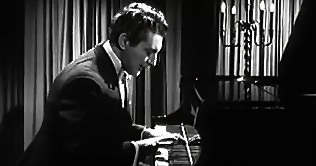 A man in a suit plays a drum set in a dimly lit room with curtains and a chandelier.