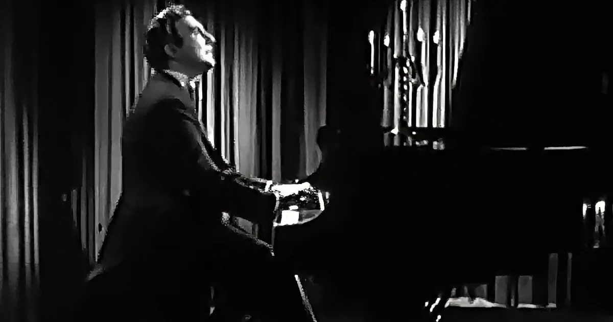 A man in a suit plays a piano in a dimly lit room with organ pipes in the background.