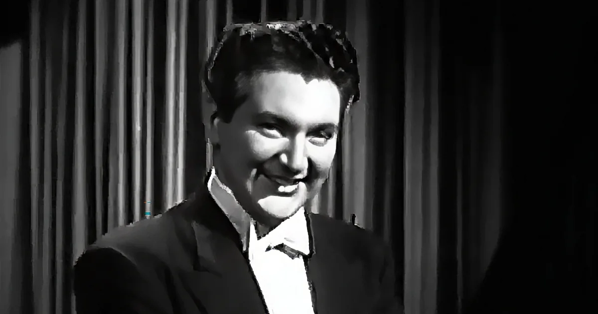 A man in a suit smiles, wearing a white shirt, against a dark, striped curtain backdrop.