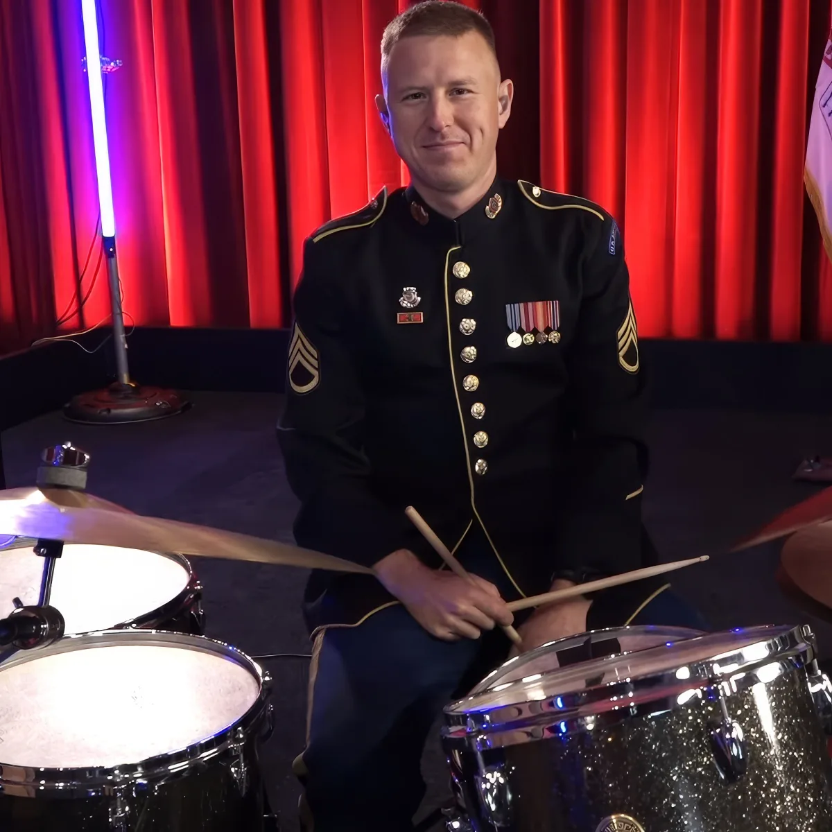 A man in a military uniform plays drums in front of red curtains.