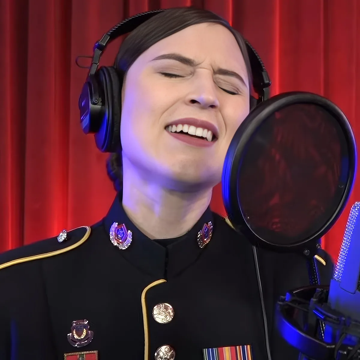 A person in a military uniform sings into a microphone with headphones, set against a red curtain backdrop.