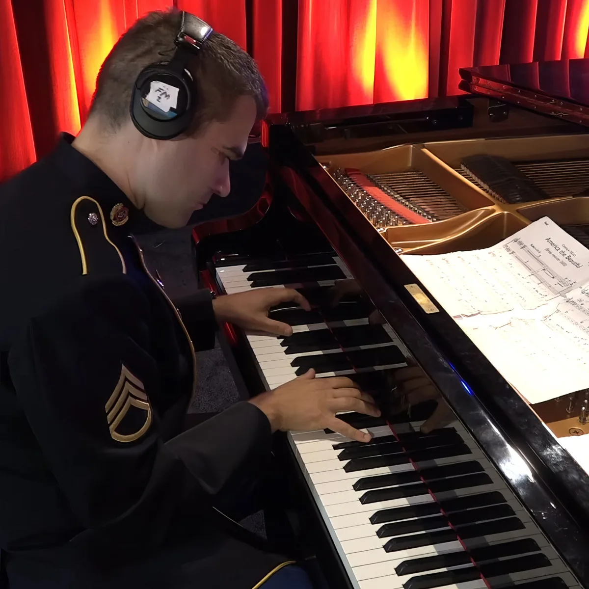 A person in uniform plays a piano with headphones, sheet music visible, red curtains in the background.