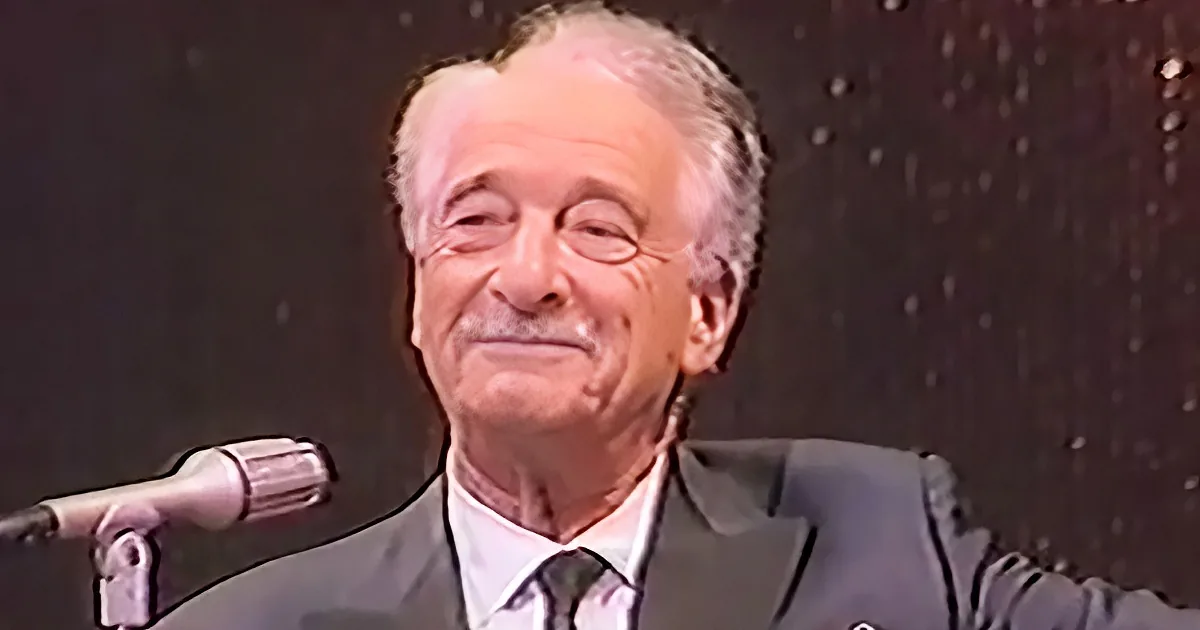 A man in a suit and tie smiles near a microphone, with a dark background.