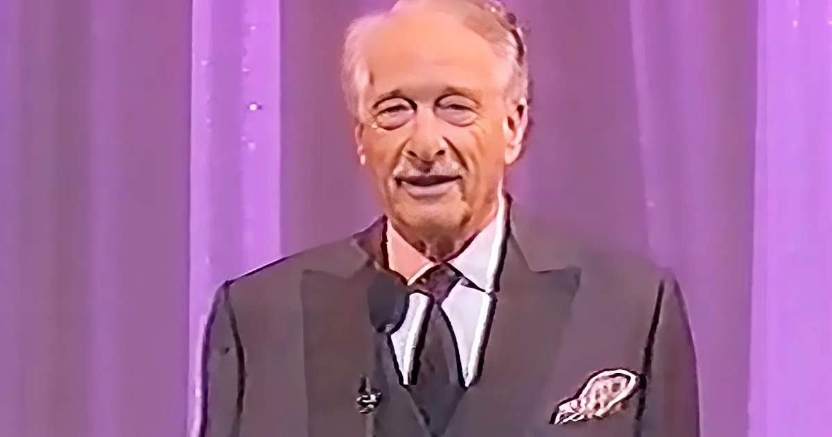 A man in a suit and tie smiles on stage with a purple backdrop.