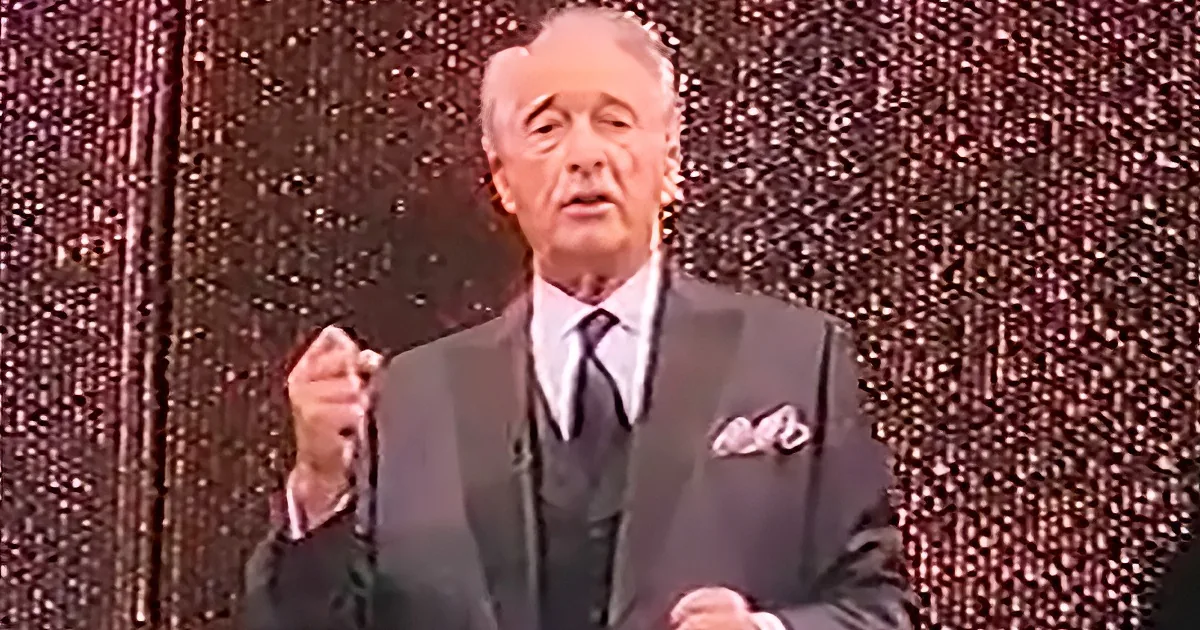 A man in a suit and tie stands against a glittery backdrop, gesturing with his right hand.