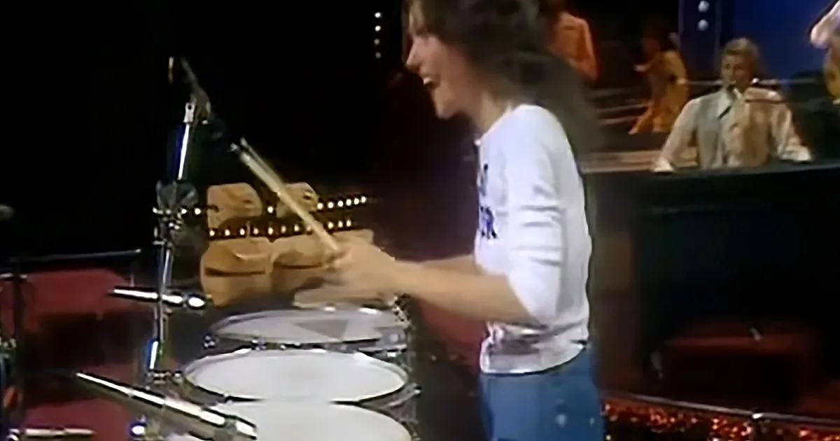 A drummer in a white shirt is playing the drums with a microphone stand nearby.