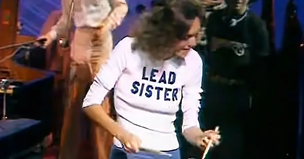 A person in a "LEAD SISTER" shirt plays drums on stage with a band.