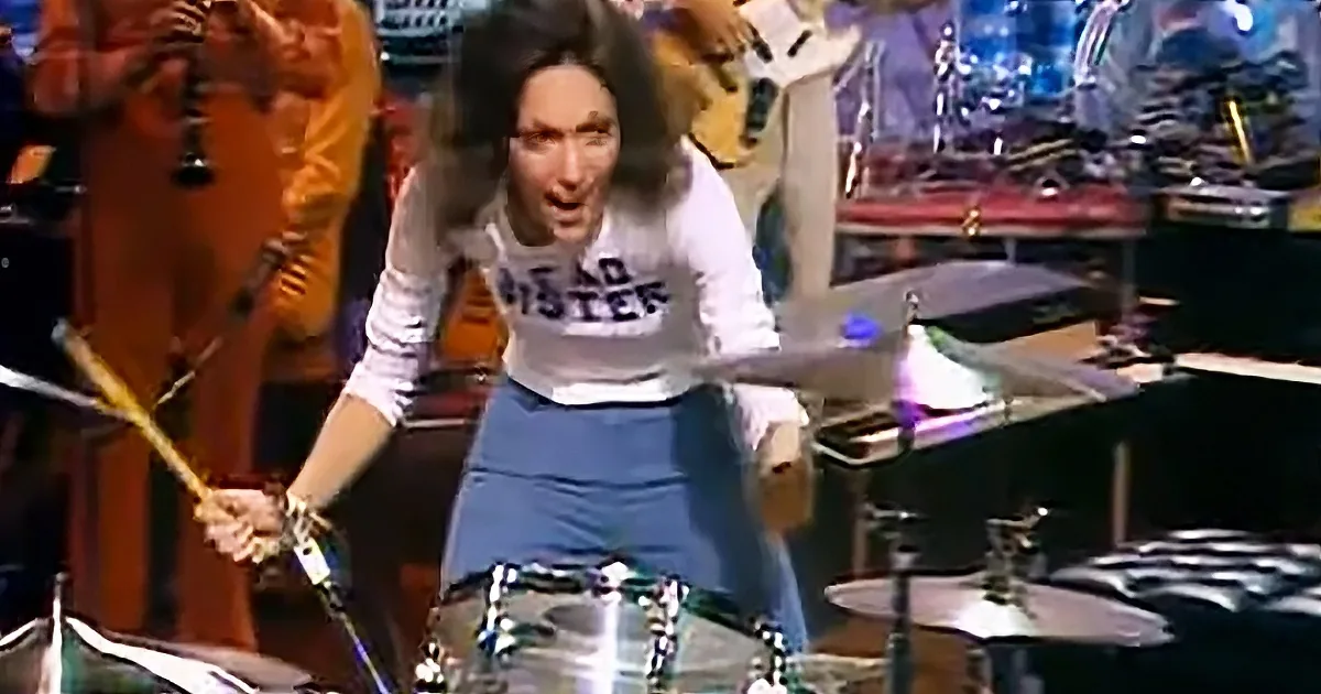 A person in a white shirt and blue skirt is playing drums on a stage with a band in the background.