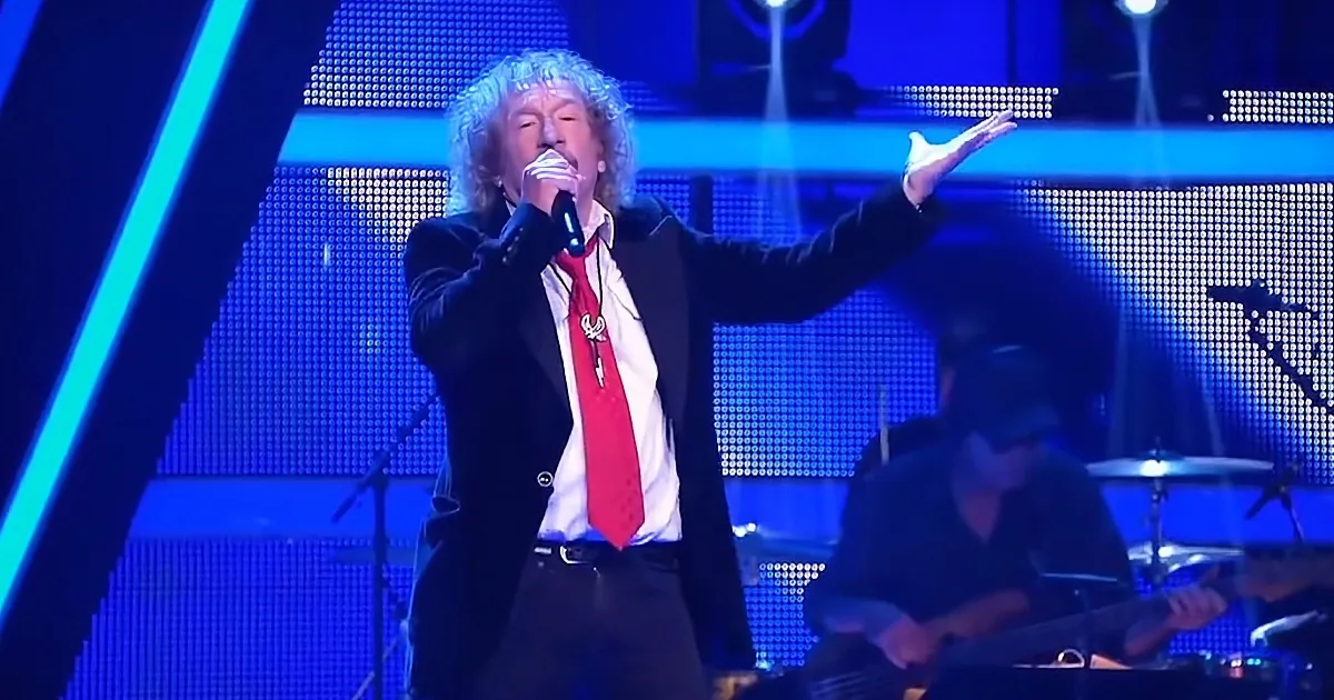 A man with curly hair sings into a microphone, wearing a red tie and black jacket, with a band playing behind him.