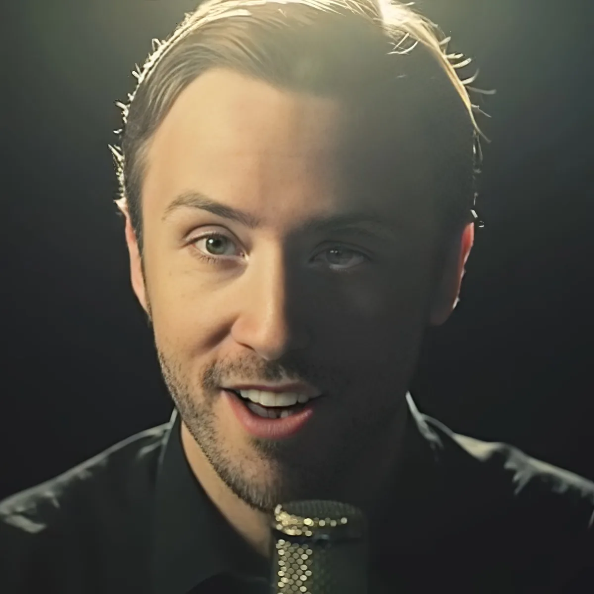 A man with short blonde hair, wearing a black shirt and a gold chain, smiles slightly in a dimly lit setting.