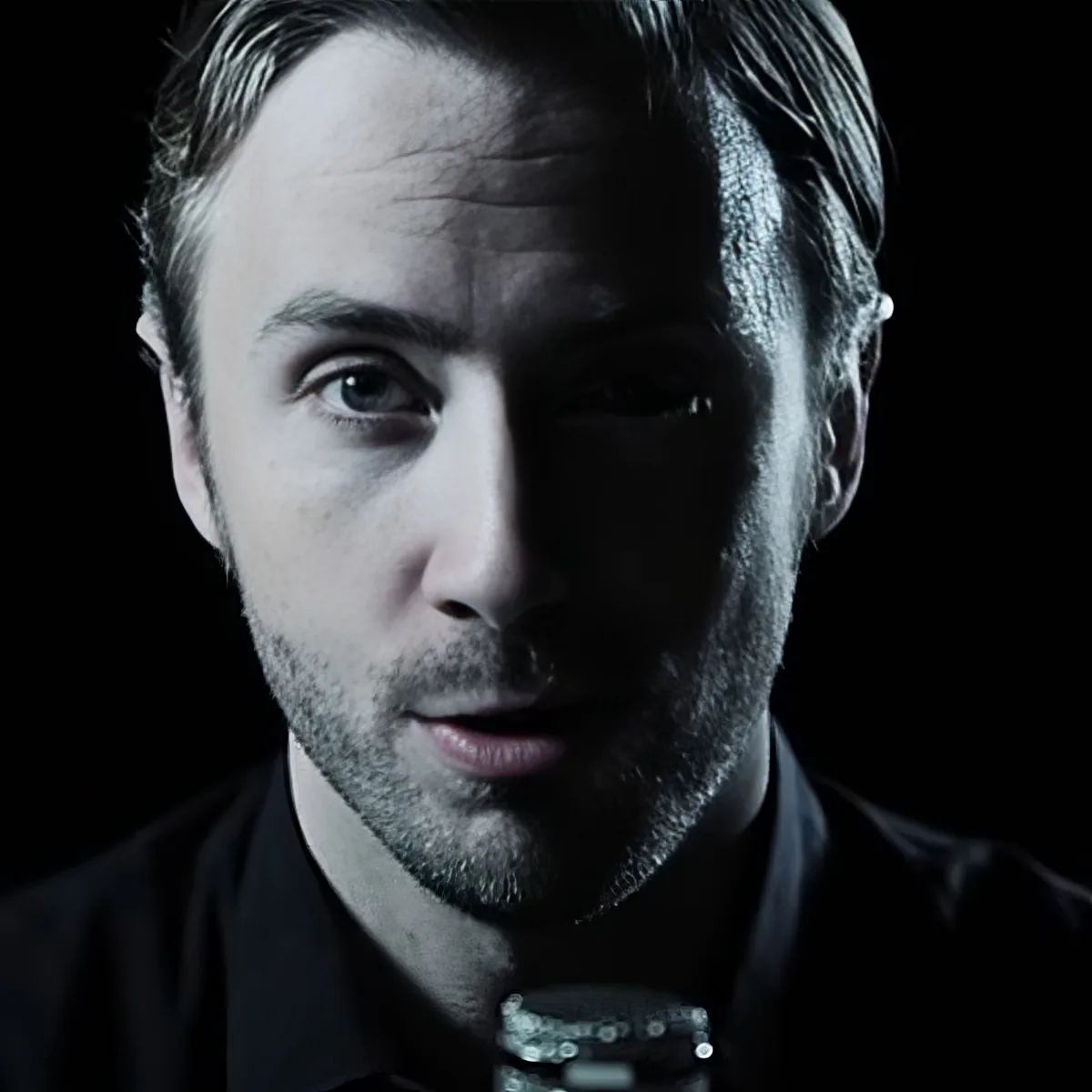 A man with short hair and a beard, wearing a dark shirt, is holding a microphone in front of his face.