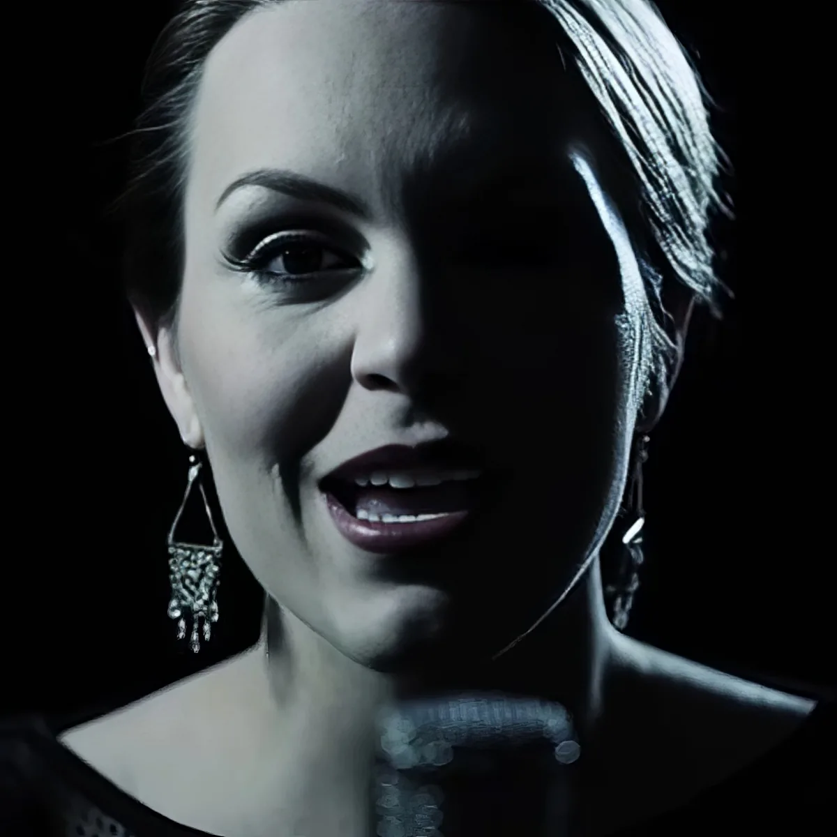 A woman with short hair, wearing dangling earrings and a dark outfit, is shown in a dimly lit setting.