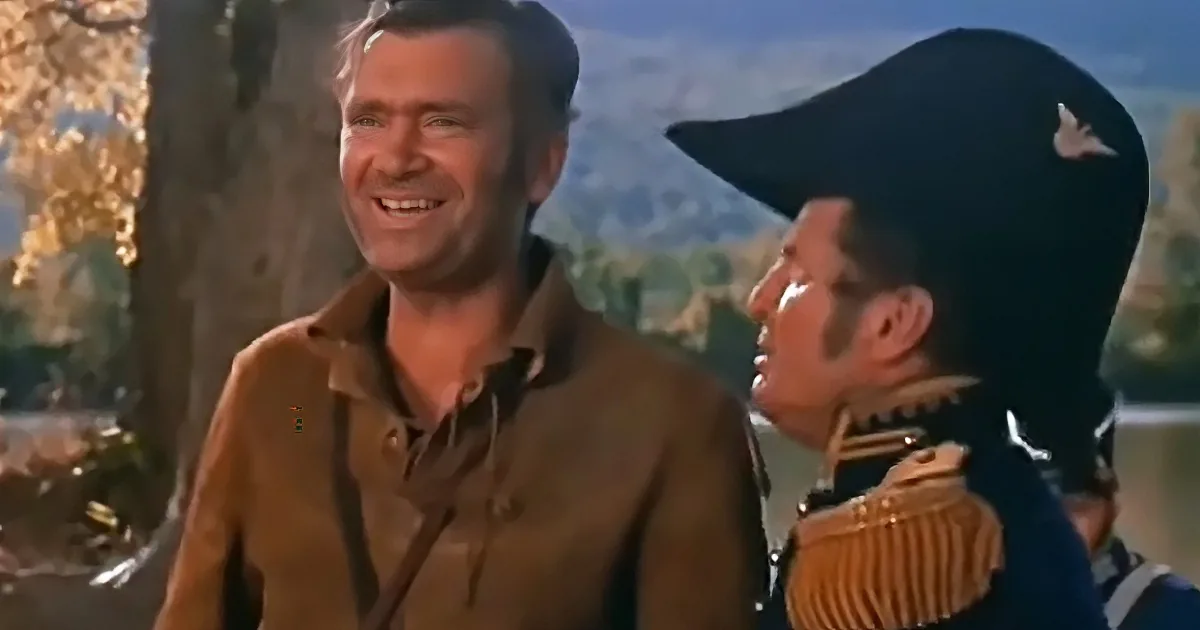 A man in a brown shirt smiles while another in a military uniform looks up, both in a scenic outdoor setting.