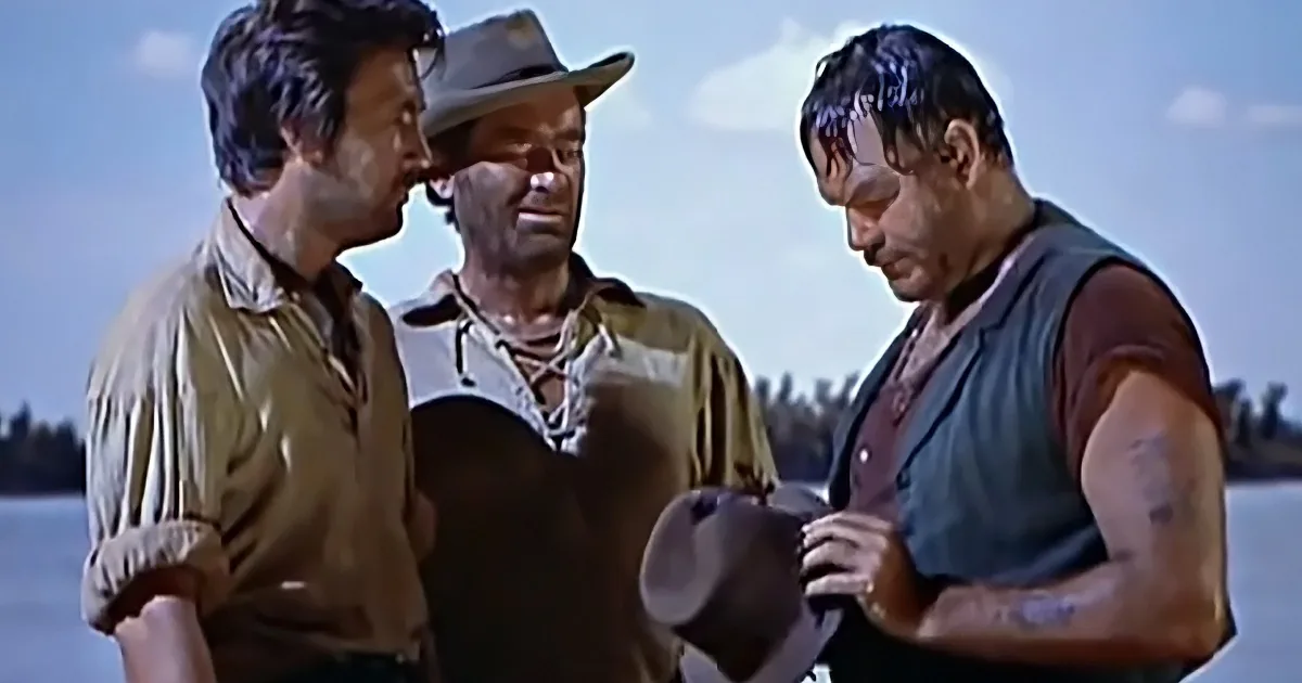 Three men in Western attire stand together, one holding a hat, against a backdrop of water and sky.