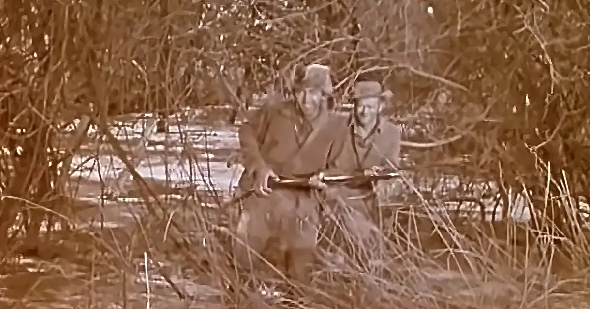 Two soldiers in camouflage gear are crouching behind a wooden barrier in a dense, dry, brushy area.