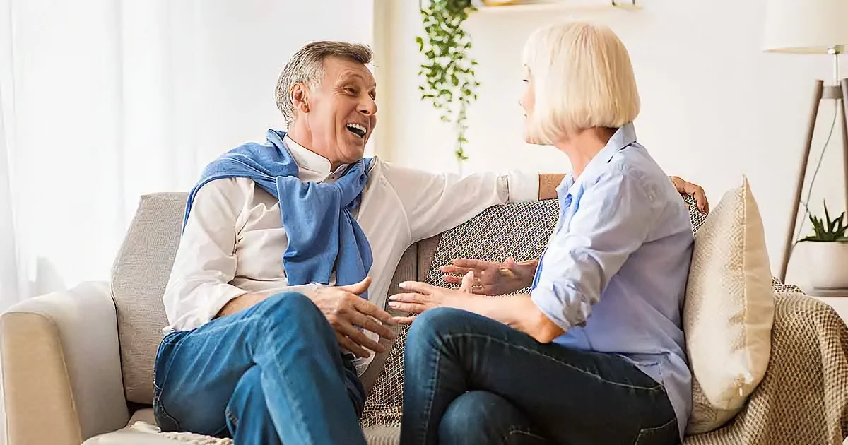 A man and woman are sitting on a couch, laughing and talking, with a plant and lamp in the background.