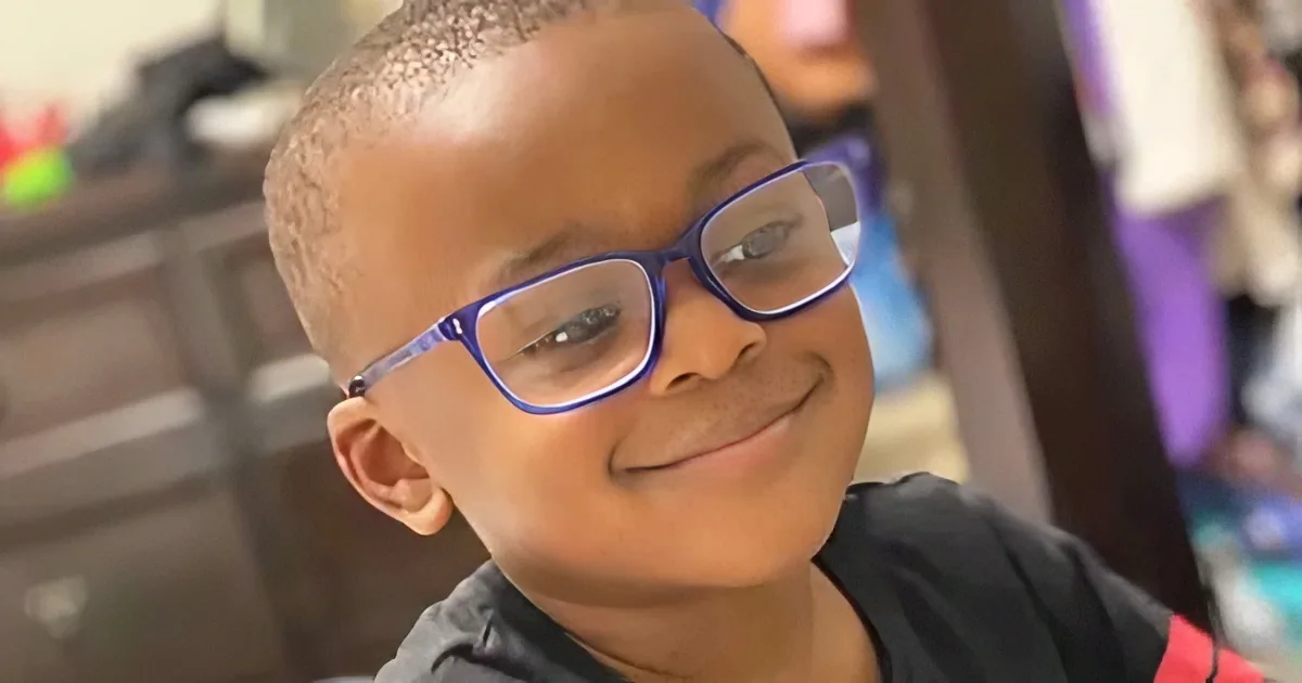 A young boy with a shaved head and glasses smiles while wearing a black shirt.
