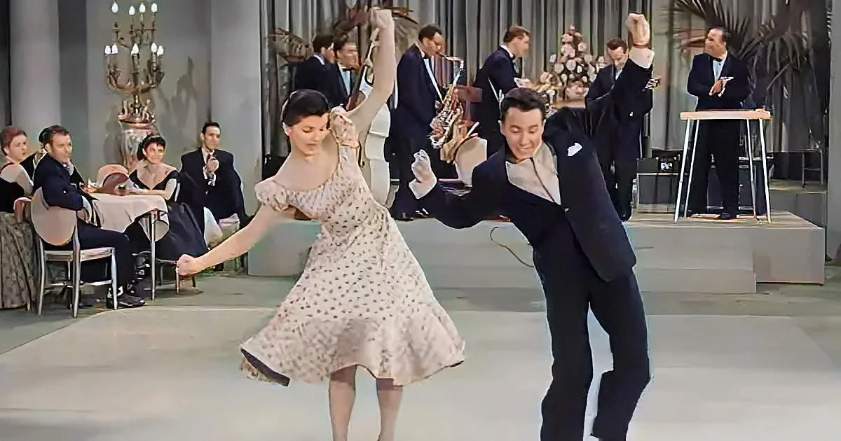 A couple dances gracefully in a formal setting, surrounded by elegantly dressed guests and a band.