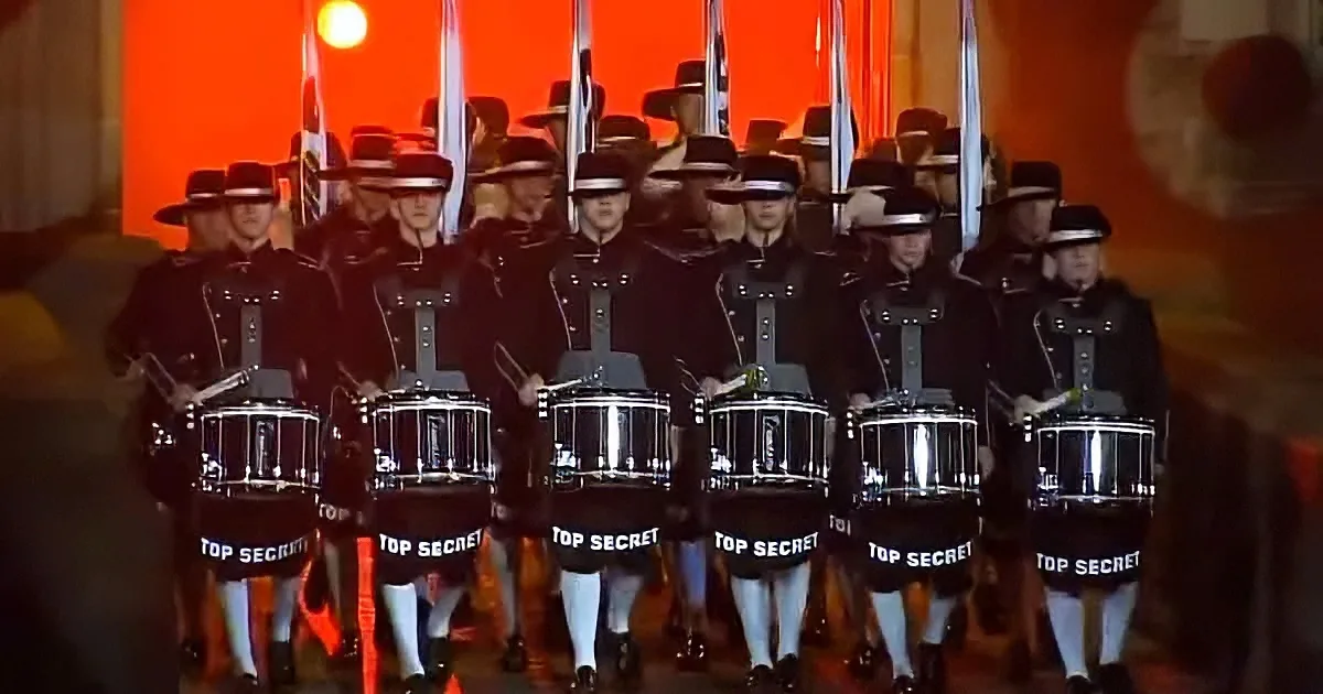 A group of drummers in uniform with "TOP SECRET" on their drum covers performs in front of a red backdrop.