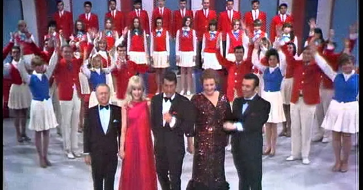A group of performers in red and white outfits, with a front row in formal attire, celebrating on stage.