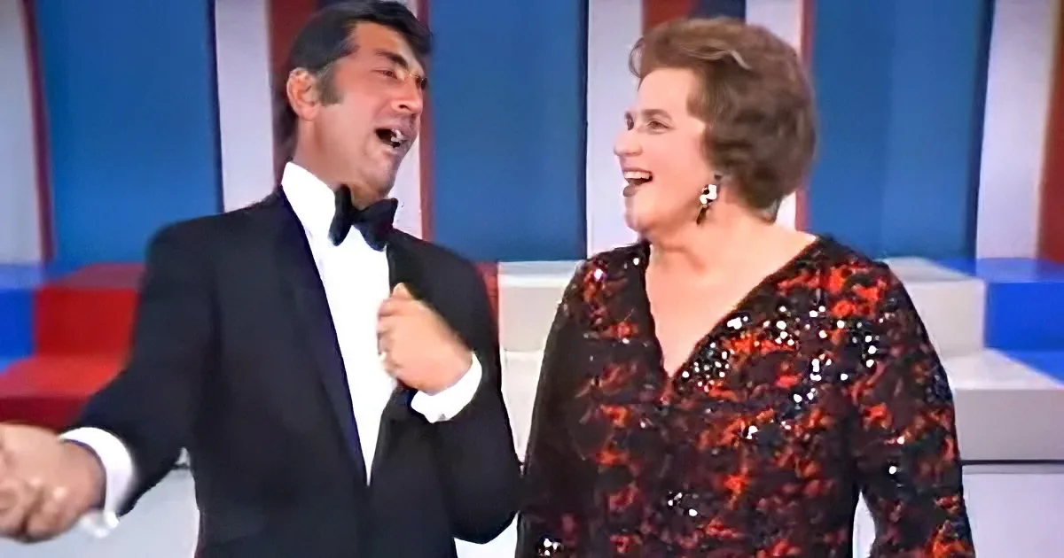 A man in a tuxedo and a woman in a sequined dress are laughing together on a stage.