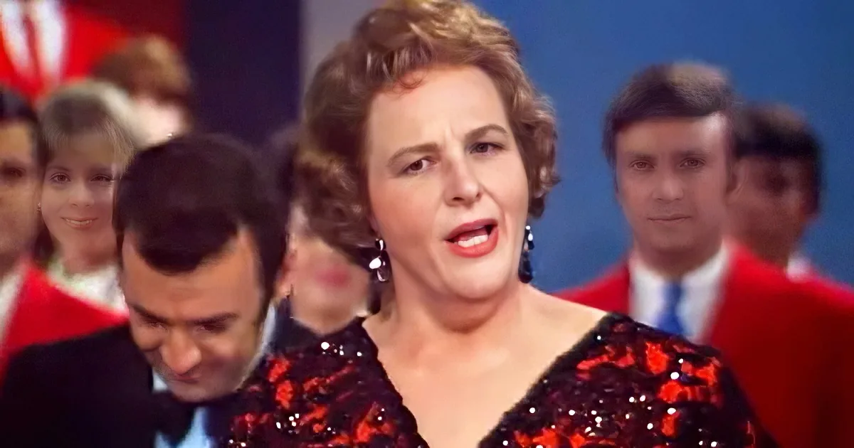 A woman in a red sequined dress sings, surrounded by a choir in red jackets and white shirts.