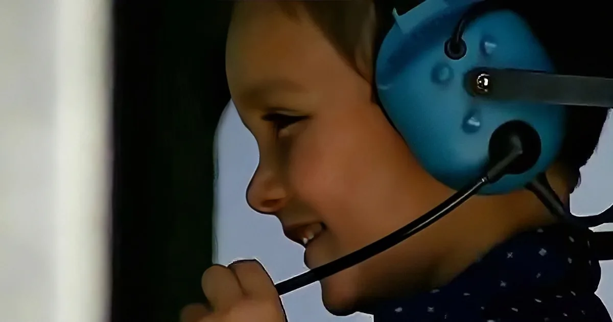 A child wearing a blue headset with a microphone is smiling and looking down.