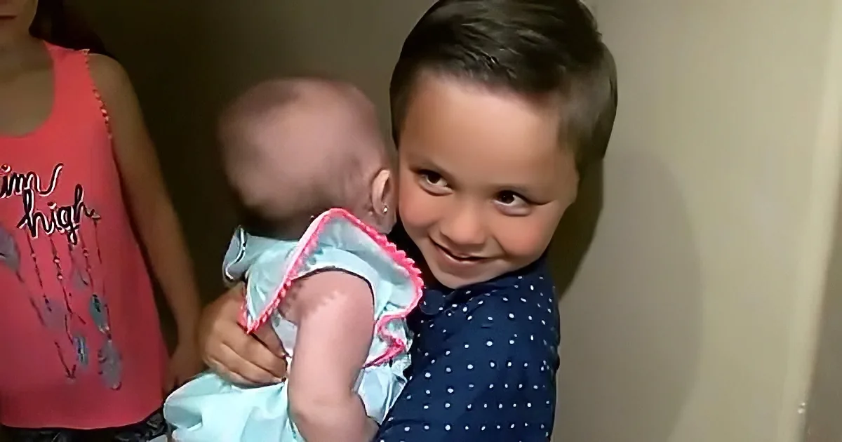 A young boy in a blue polka-dot shirt holds a baby in a blue dress, smiling warmly.