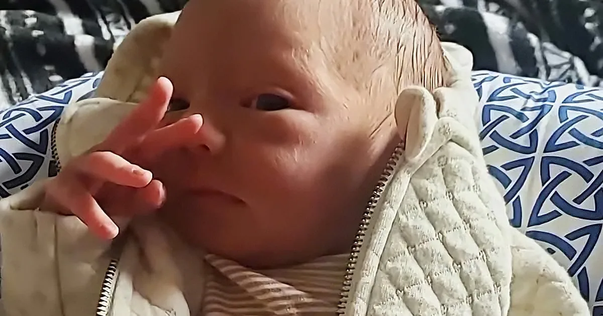 A baby wearing a quilted jacket rests on a patterned surface, holding its hand near its face.