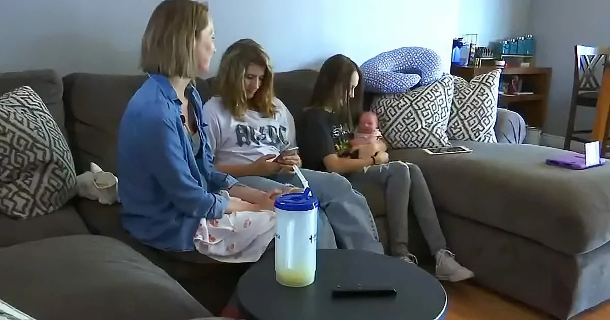 Three girls sit on a couch, one holding a baby, another using a phone, and the third looking down.