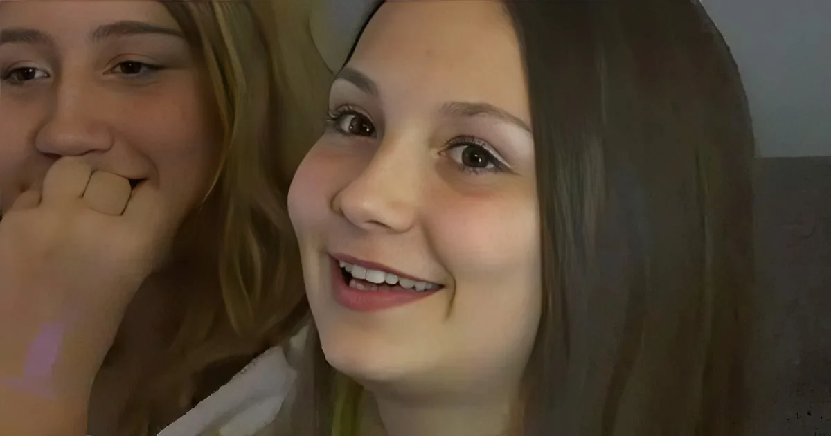 A young girl with long brown hair smiles, showing a missing tooth, while another person in the background looks on.