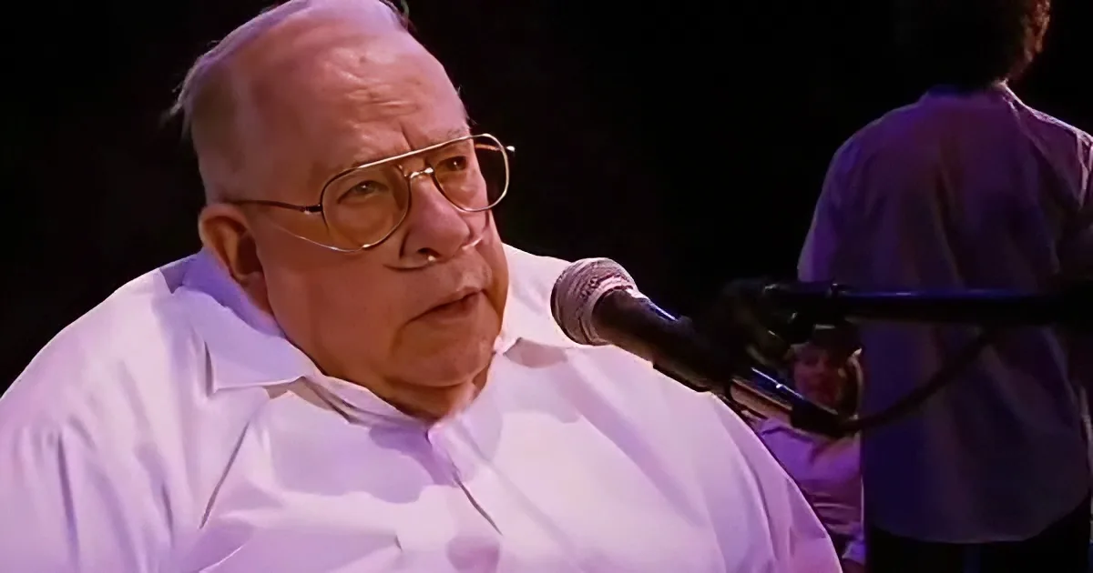 An older man in a white shirt speaks into a microphone, wearing glasses, with a dark background and another person...