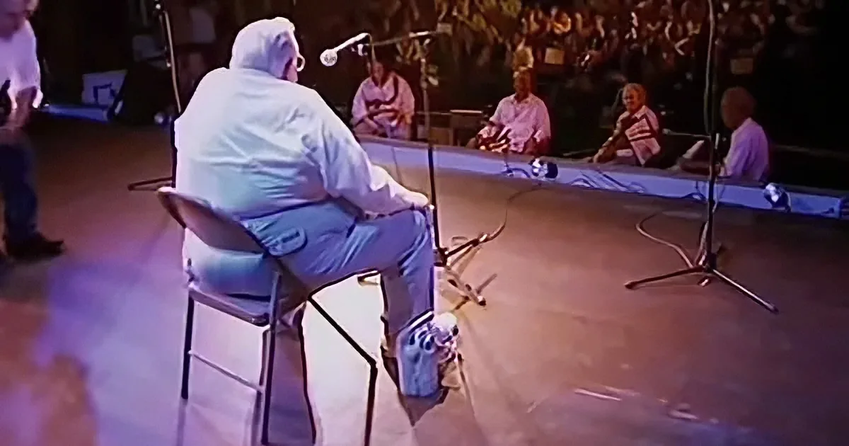 A person in a white jumpsuit sits on a chair, facing a microphone on a stage with other people in the background.