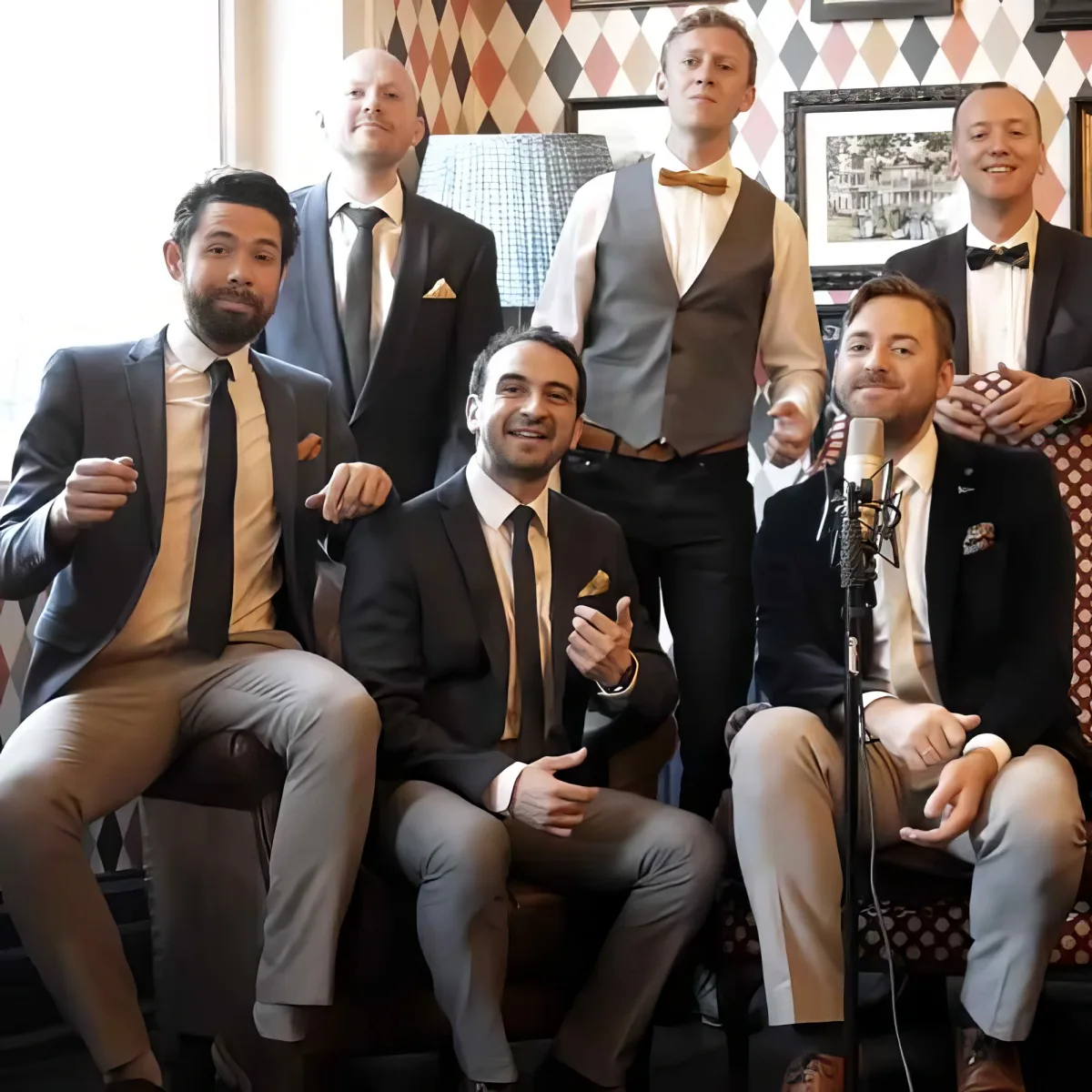 A group of men in formal attire, some seated and one holding a microphone, in a room with patterned walls.
