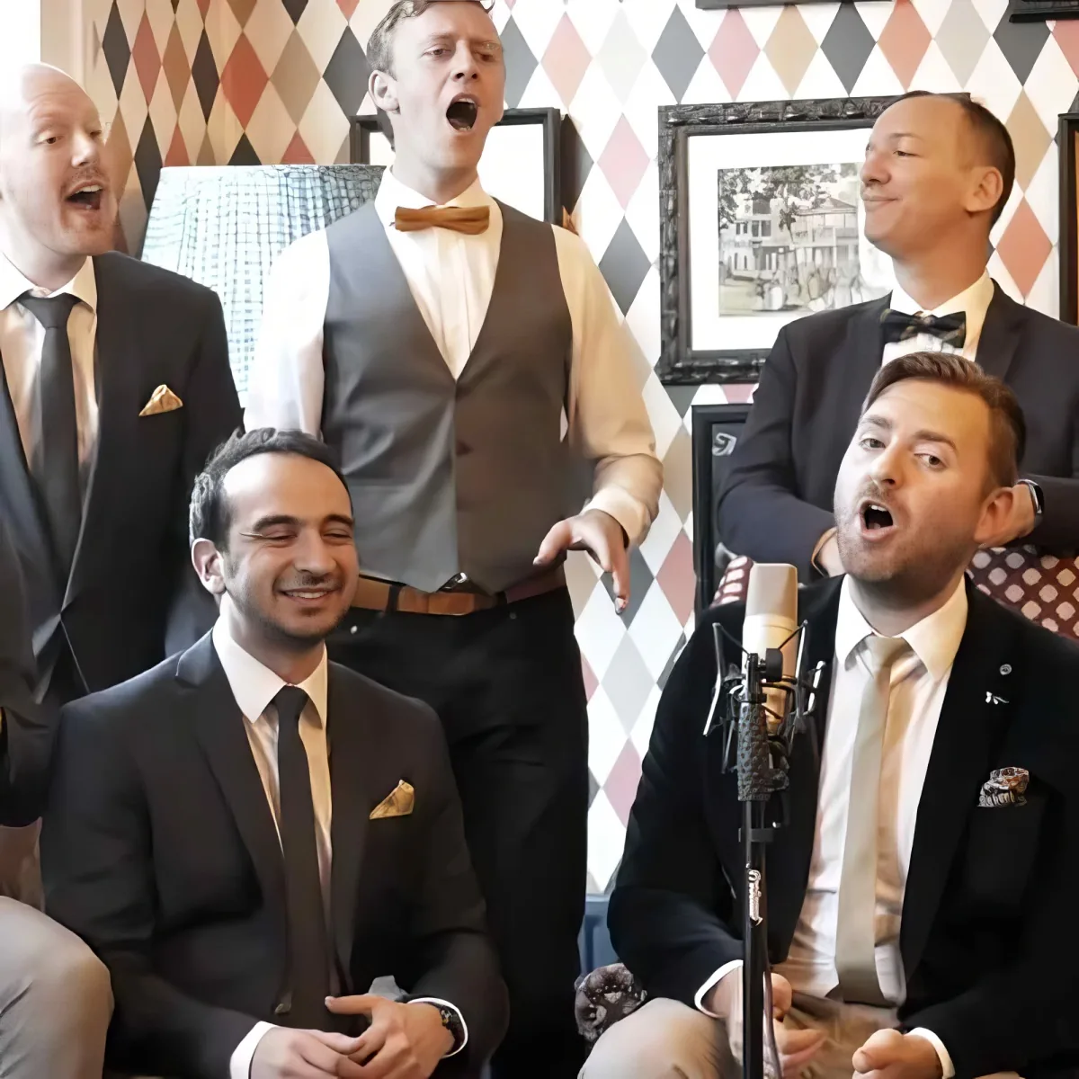 A group of men in suits and ties sing and smile in front of a harlequin-patterned wall.
