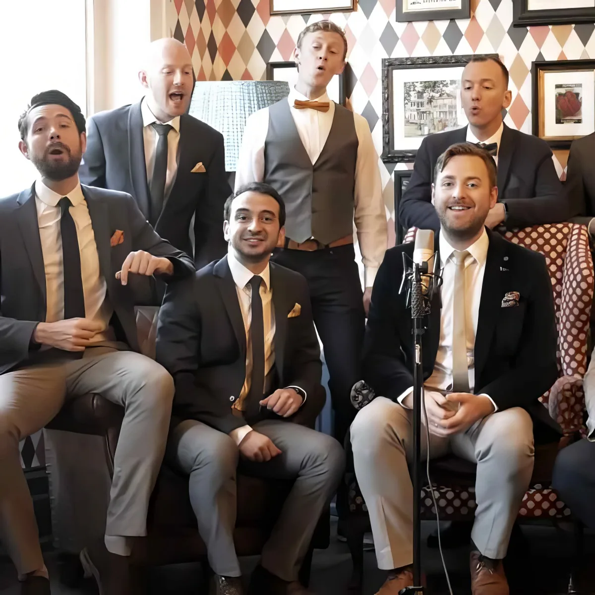A man in a suit sits with a microphone, surrounded by a group of men in suits, some singing.