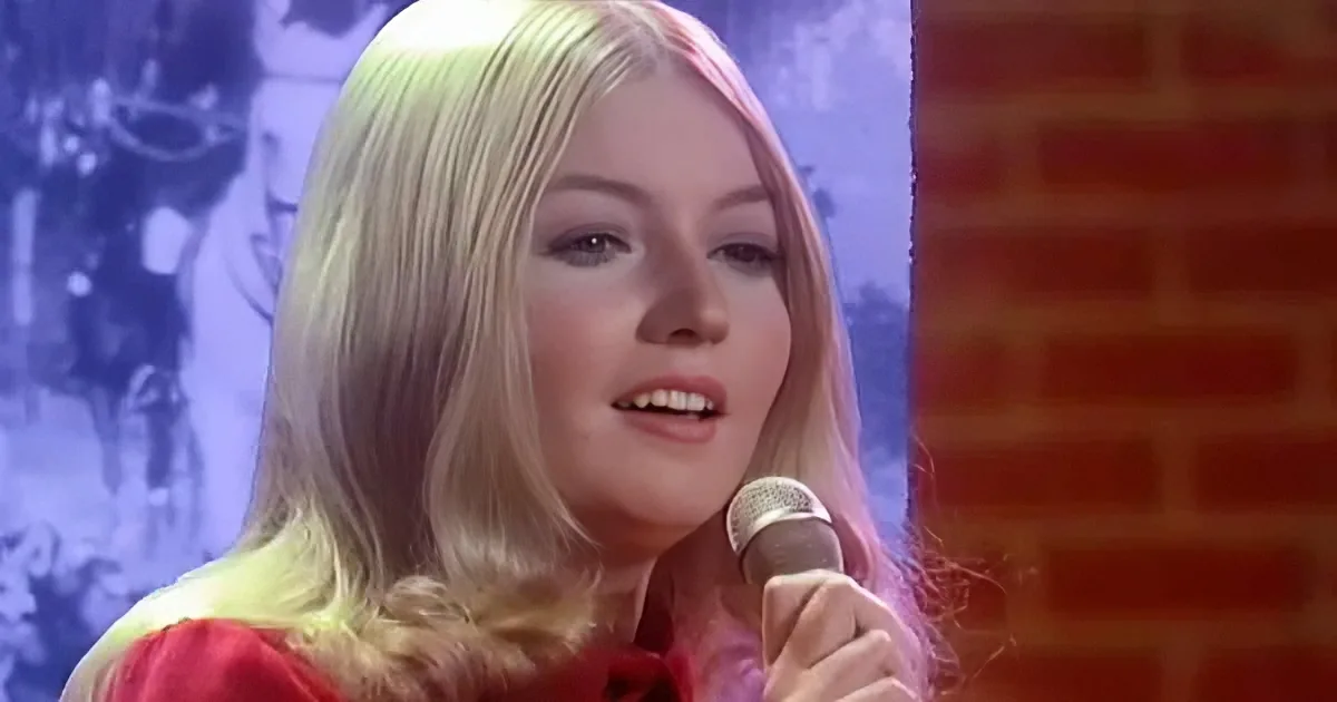 A person with long blonde hair, wearing a red outfit, holds a microphone, possibly singing or speaking.