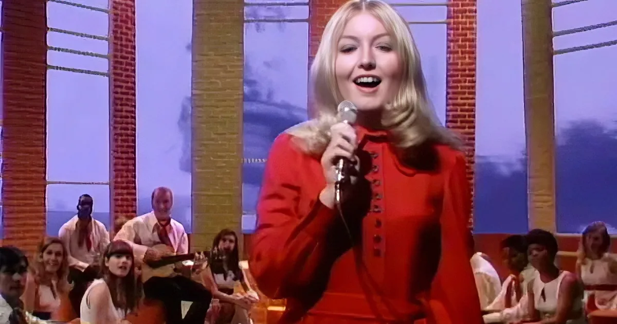 A woman in a red dress sings into a microphone on stage, with musicians and an audience in the background.