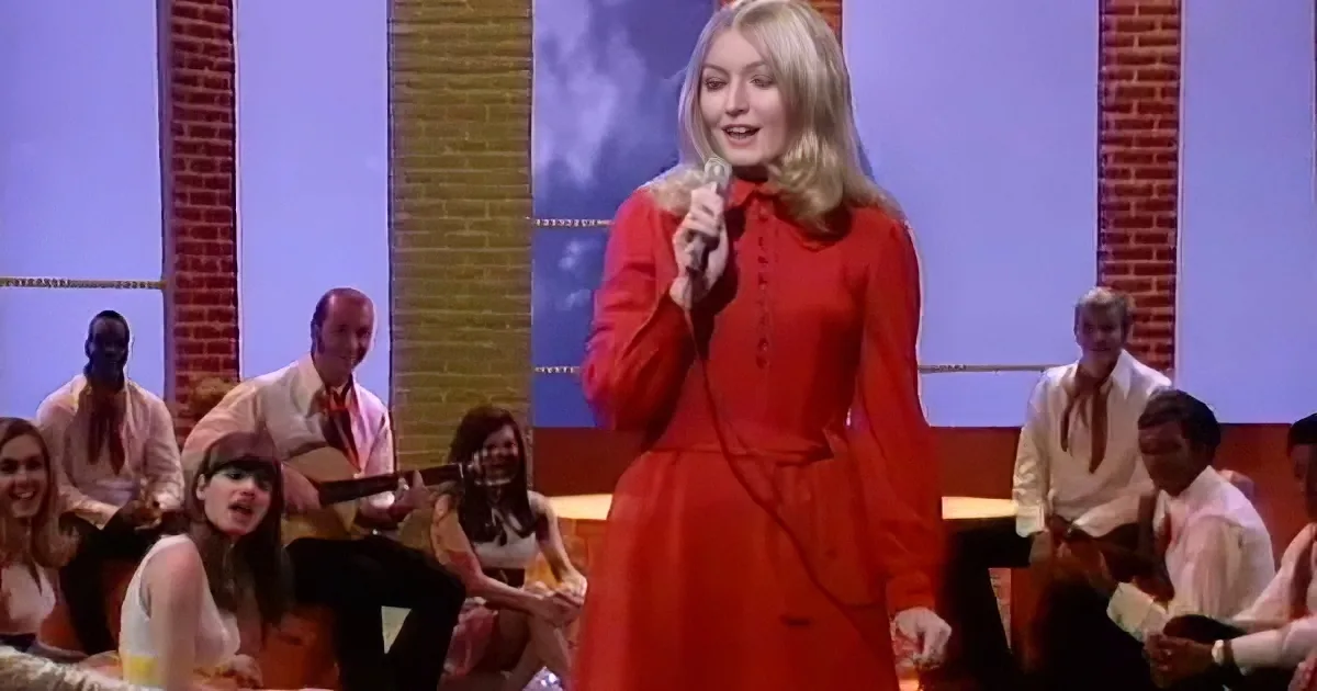 A woman in a red dress sings into a microphone on stage with musicians and a brick backdrop.