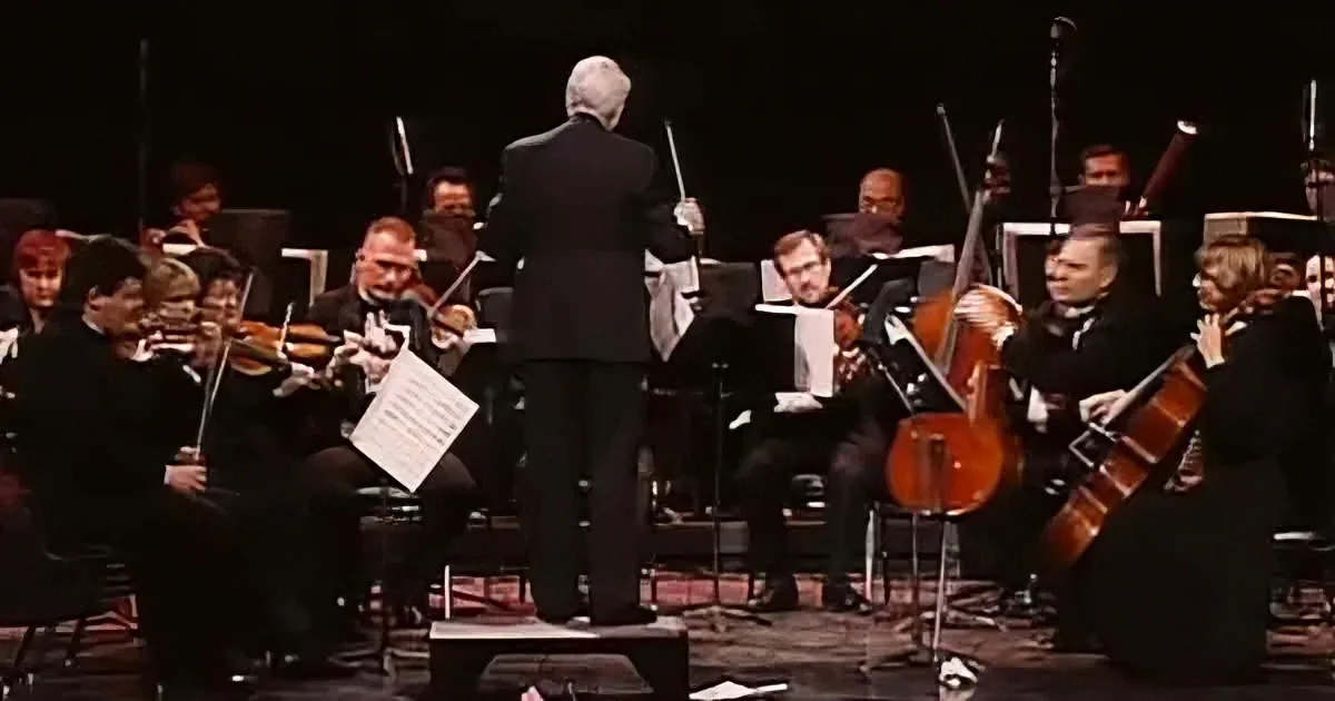 Conductor leads orchestra with violinists and cellists in formal attire on stage.