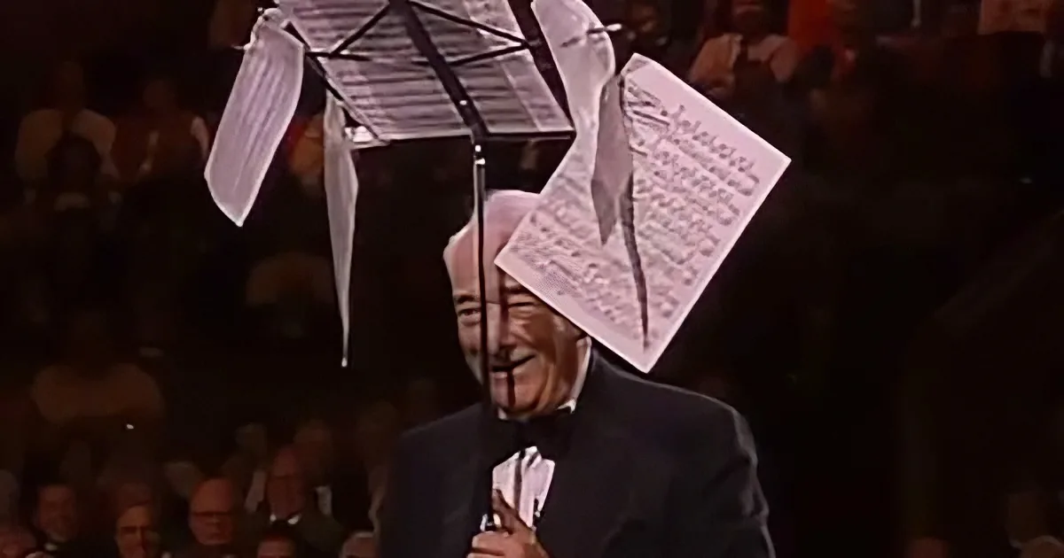An elderly man in a tuxedo smiles as sheet music hangs from a music stand above him.