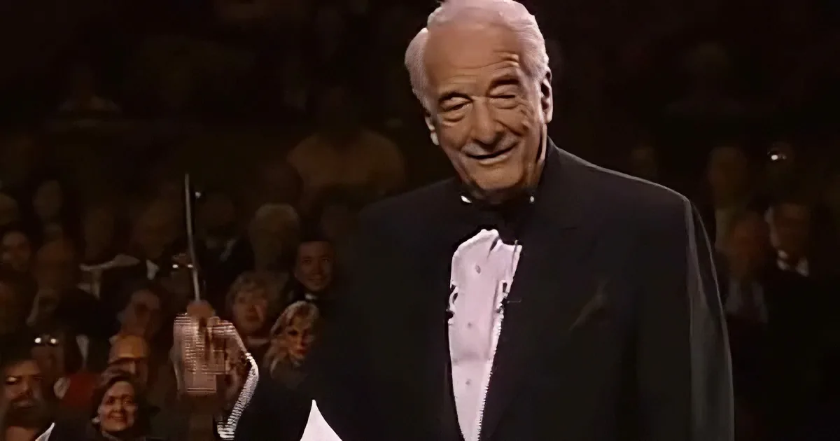 An older man in a tuxedo smiles while holding a microphone, with a crowd in the background.