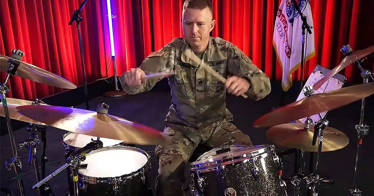 A soldier in camouflage plays drums on a stage with red curtains and a flag.